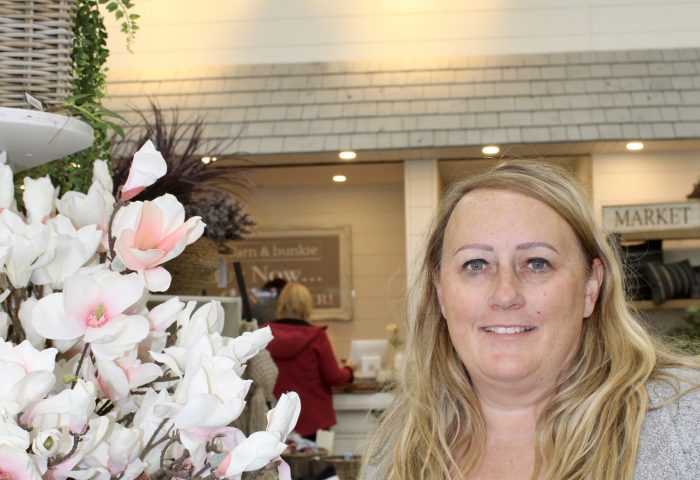 A woman stands beside flowers. On the wall behind her, are the words "barn and bunkie".