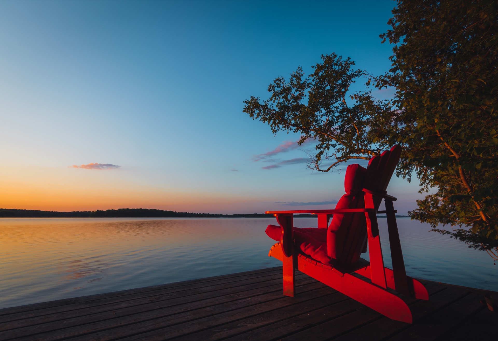 Two red Muskoka chairs on a dock overlooking a Kawartha lake at sunset