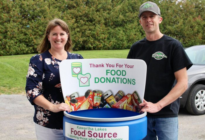 Two people smiling standing beside a food donation barrel